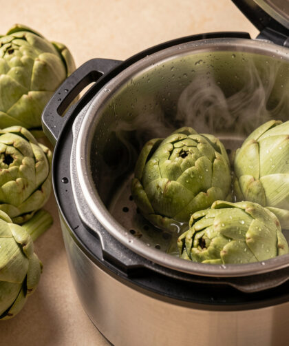 Artichauts de différentes tailles disposés autour d’une cocotte-minute en acier inoxydable, avec de la vapeur visible pendant la cuisson.